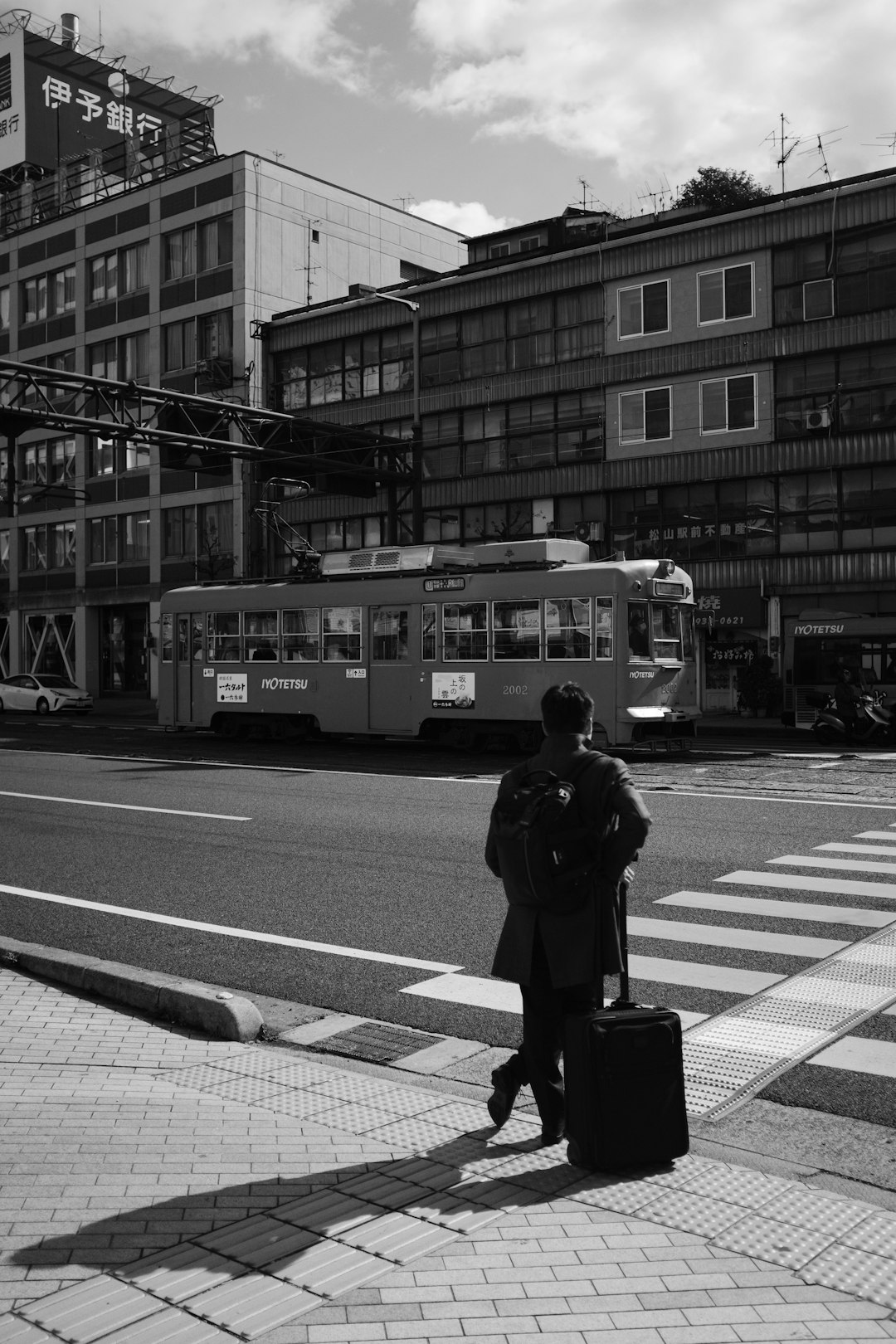 grayscale photo of man in black jacket and black pants carrying black luggage bag walking on near near near near