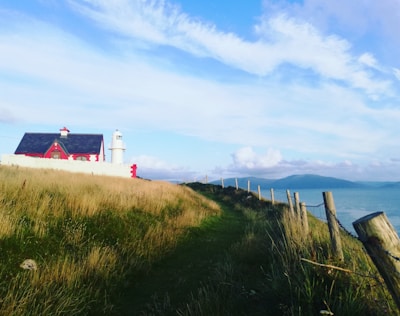 A scenic coastal view featuring a red and white lighthouse nestled beside a small house. The scene is bordered by a grassy path and a rustic wooden fence leading towards the sea on the right. The backdrop consists of a clear blue sky with scattered clouds and distant hills across the water.