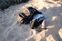 A black backpack, a blue denim shirt, a black water bottle, and a pink portable speaker are positioned on a sunlit sandy beach. The sand is slightly indented where the objects are placed, and there are sparse patches of vegetation in the background.