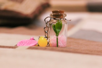 A small glass bottle with a cork stopper contains a green plant stem and pink granular material. Attached to the bottle is a metal chain with a yellow and pink charm.