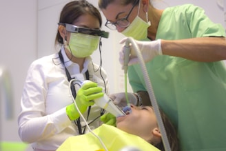 Two dental professionals perform a procedure on a patient. One is wearing specialized eyewear and is focused on using a dental instrument. The patient lies in a dental chair, wearing a protective bib.