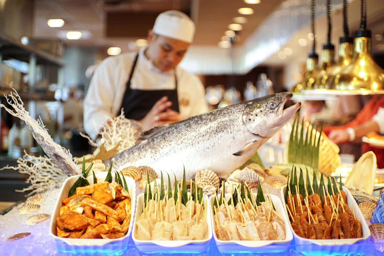 A chef in a white uniform preparing a whole salmon at a seafood buffet with skewers and prepared dishes