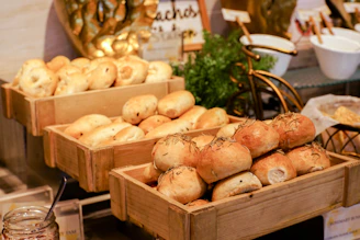 An assortment of freshly baked bread rolls is displayed in wooden crates. The rolls are golden brown with some topped with fresh herbs. The setting suggests a buffet or bakery with an inviting atmosphere. In the background, there are decorative elements including a small bicycle and some greenery.