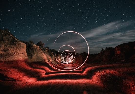 A night sky filled with stars forms the background, while a rocky desert landscape is illuminated by red lights. Intricate spirals of light dominate the center, creating a mesmerizing trail along the rugged terrain. Clouds add texture to the sky, enhancing the overall mystique.