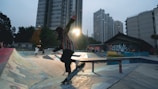 A skateboarder performing a trick on a ramp in an urban skate park. The skate park features graffiti art on the walls and ramps, and the scene is set in an urban area with tall buildings in the background. The lighting suggests it is either dawn or dusk, giving the scene a moody atmosphere.