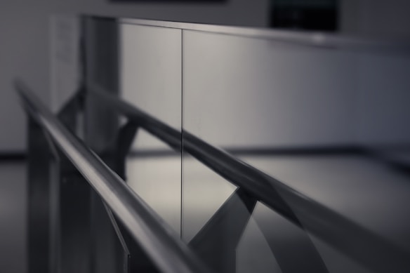A close-up perspective of a metallic handrail with reflective glass panels, creating a symmetrical composition reminiscent of modern architecture. The image has a shallow depth of field, focusing on the left side while the background is blurred, emphasizing the sleek and clean design. The lighting is soft, adding a subtle shine to the reflective surfaces.