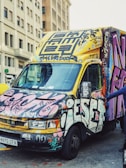A van parked on a city street is covered in colorful graffiti and street art. The vehicle features a mixture of abstract designs and lettering in vibrant shades of pink, yellow, blue, and black. Surrounding buildings have a classic architectural style with multiple windows.
