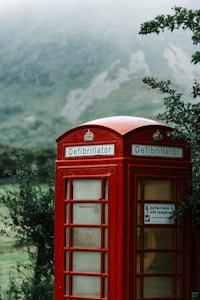 A red defibrillator box designed like a traditional British telephone booth stands in a lush, green rural setting with mountains and mist in the background.