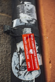 A weathered, textured black pipe is affixed to a wall with peeling posters partially covering its surface. A vibrant red sticker with white text in German reading 'KEIN LEBEN OHNE FREIHEIT' alongside 'FIGHT CAPITALISM' and '100%' is prominently displayed.