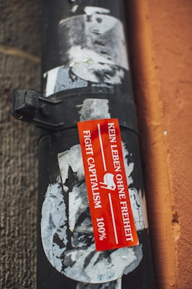 A weathered, textured black pipe is affixed to a wall with peeling posters partially covering its surface. A vibrant red sticker with white text in German reading 'KEIN LEBEN OHNE FREIHEIT' alongside 'FIGHT CAPITALISM' and '100%' is prominently displayed.