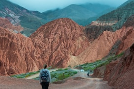 A person with a backpack walks along a dirt path surrounded by striking, rugged red rock formations and green vegetation. In the distance, there are two more people walking and a white car parked on the side of the path. The scene is set against a backdrop of misty, blue-tinted mountains.