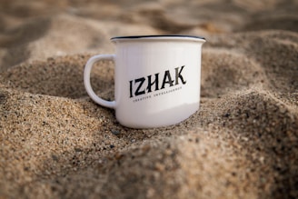 A white mug with the text 'IZHAK Creative Intelligence' printed on it is placed in a setting of sand. The mug is positioned upright, surrounded by grainy sand, and it appears to be a calm and serene environment.