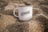 A white mug with the text 'IZHAK Creative Intelligence' printed on it is placed in a setting of sand. The mug is positioned upright, surrounded by grainy sand, and it appears to be a calm and serene environment.