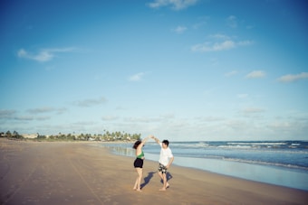 A couple dances joyfully on a vast, nearly empty beach under a bright blue sky. The shoreline stretches far into the distance with gentle waves lapping at the sand. In the background, palm trees and a few buildings are visible.
