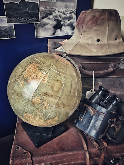 A vintage scene featuring a large globe on a stack of old leather suitcases. There is an explorer hat placed on top, alongside a pair of binoculars in a worn leather case. Black and white photographs of landscapes are displayed on the wall in the background.
