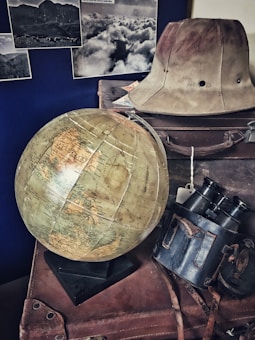 A vintage scene featuring a large globe on a stack of old leather suitcases. There is an explorer hat placed on top, alongside a pair of binoculars in a worn leather case. Black and white photographs of landscapes are displayed on the wall in the background.