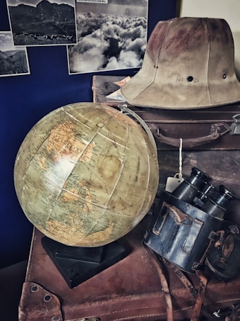 A vintage scene featuring a large globe on a stack of old leather suitcases. There is an explorer hat placed on top, alongside a pair of binoculars in a worn leather case. Black and white photographs of landscapes are displayed on the wall in the background.
