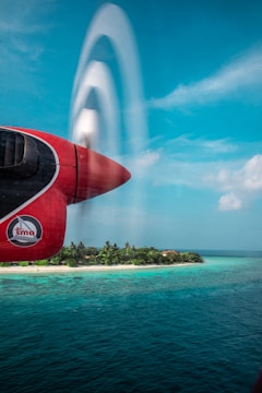 A red airplane engine is in the foreground, with its propeller blades creating a blurred, translucent effect against a bright blue sky. Below, a tropical island with lush green palm trees and a sandy beach is surrounded by vibrant turquoise water.