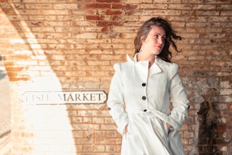 A person in a white coat stands against a brick wall with a sign that reads 'FISH MARKET.' The person has wavy hair slightly blown by the wind, looking off to the side, with a relaxed and contemplative expression.