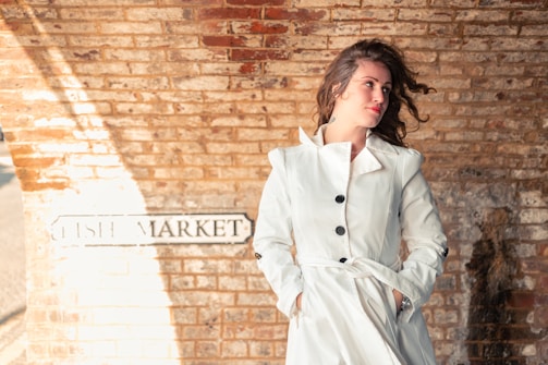 A person in a white coat stands against a brick wall with a sign that reads 'FISH MARKET.' The person has wavy hair slightly blown by the wind, looking off to the side, with a relaxed and contemplative expression.