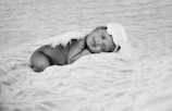 A newborn baby is peacefully sleeping on a soft, textured blanket wearing a headband adorned with a large flower and angelic feather wings.