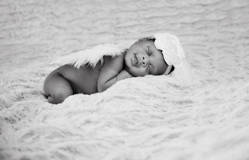 A newborn baby is peacefully sleeping on a soft, textured blanket wearing a headband adorned with a large flower and angelic feather wings.