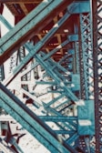 A close-up view of the intricate metal framework of a bridge, showcasing a complex pattern of blue steel beams and rivets. The structure highlights geometric lines and angles, creating a visually striking industrial scene.