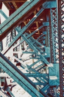 A close-up view of the intricate metal framework of a bridge, showcasing a complex pattern of blue steel beams and rivets. The structure highlights geometric lines and angles, creating a visually striking industrial scene.
