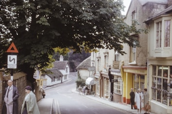 A quaint, tree-lined street scene in a small village, featuring brick buildings with charming shops and a thatched-roof cottage in the background. Several people dressed in coats are walking or standing along the sidewalk. A traffic sign indicating a double bend is visible, and a bicycle is parked nearby. The scene has a vintage and serene atmosphere, enhanced by the overcast lighting.