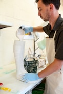 A person wearing a white apron and blue gloves pours a white liquid substance from a measuring cup into a tall boot-shaped mold. The setting appears to be an art or craft studio, with other tools and materials visible in the background.
