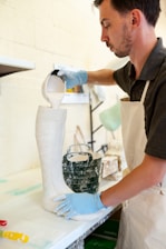 A person wearing a white apron and blue gloves pours a white liquid substance from a measuring cup into a tall boot-shaped mold. The setting appears to be an art or craft studio, with other tools and materials visible in the background.