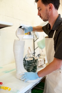 A person wearing a white apron and blue gloves pours a white liquid substance from a measuring cup into a tall boot-shaped mold. The setting appears to be an art or craft studio, with other tools and materials visible in the background.
