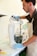 A person wearing a white apron and blue gloves pours a white liquid substance from a measuring cup into a tall boot-shaped mold. The setting appears to be an art or craft studio, with other tools and materials visible in the background.