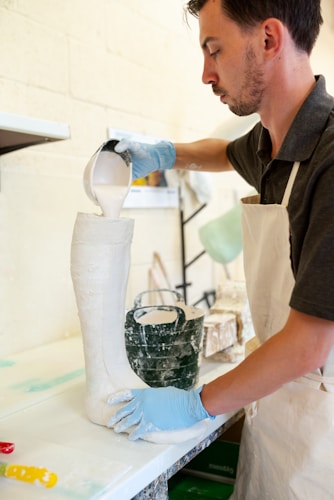 A person wearing a white apron and blue gloves pours a white liquid substance from a measuring cup into a tall boot-shaped mold. The setting appears to be an art or craft studio, with other tools and materials visible in the background.