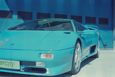 A sleek blue sports car is parked outside a building with a blue wall featuring several small windows. The car has a license plate with the word 'supercity' on it, and its aerodynamic design includes elements like a spoiler and large wheels.