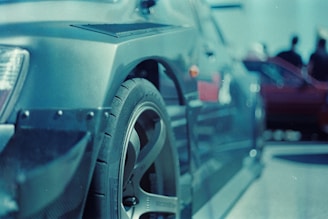 A close-up of a car wheel and part of the side body, showcasing the intricate details of the tire tread and alloy rim. The scene has a blurred background with shapes that suggest other cars and people, indicating a gathering or car show environment.