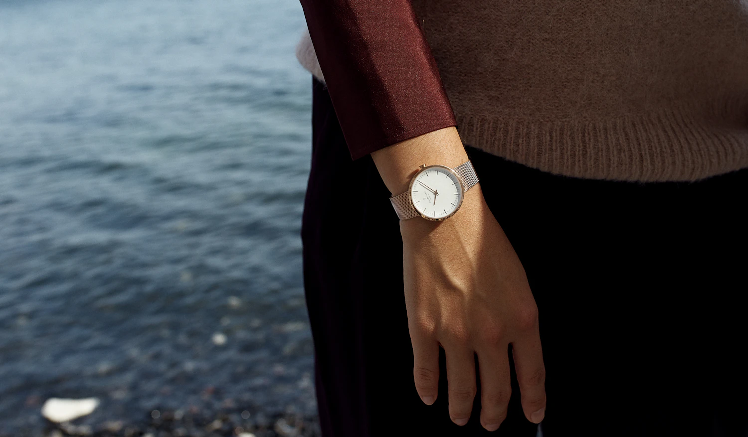 A woman by the lake wearing a small white-dial watch on a mesh bracelet