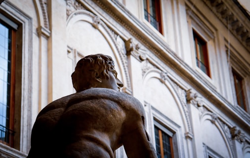 A stone sculpture depicting a muscular male figure is prominently featured in the foreground, focusing on the back and shoulder muscles. The background includes an ornate, old-style building with large windows and intricate architectural details.
