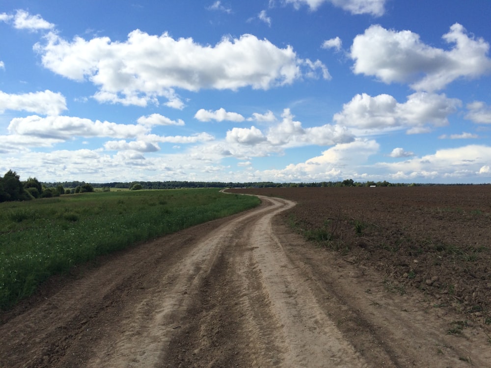brown dirt road between green grass field under blue and white cloudy sky during daytime
