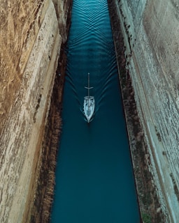 Aerial view of a sailboat navigating through a narrow canal with steep rock walls on either side. The water is a deep turquoise color, and the boat creates ripples as it moves forward.