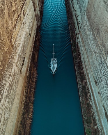 Aerial view of a sailboat navigating through a narrow canal with steep rock walls on either side. The water is a deep turquoise color, and the boat creates ripples as it moves forward.