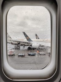 Raindrops cover an airplane window, through which a parked airplane with visible engines and a partially obscured emblem on the tail is seen on the wet tarmac. Ground handling equipment and a person in high-visibility clothing are visible near the aircraft.