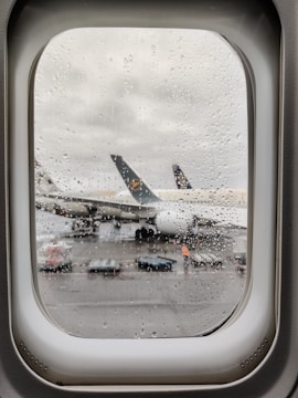 Raindrops cover an airplane window, through which a parked airplane with visible engines and a partially obscured emblem on the tail is seen on the wet tarmac. Ground handling equipment and a person in high-visibility clothing are visible near the aircraft.