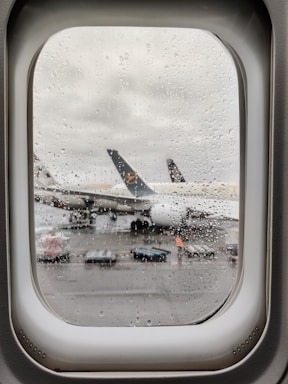 Raindrops cover an airplane window, through which a parked airplane with visible engines and a partially obscured emblem on the tail is seen on the wet tarmac. Ground handling equipment and a person in high-visibility clothing are visible near the aircraft.