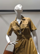 A white mannequin wearing a belted, short-sleeved, golden brown dress with button closures and pockets. It holds a tan handbag and features a colorful necklace.