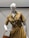 A white mannequin wearing a belted, short-sleeved, golden brown dress with button closures and pockets. It holds a tan handbag and features a colorful necklace.