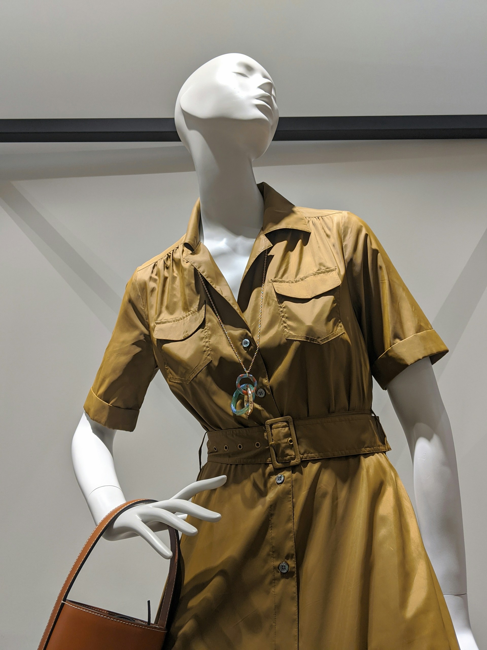 A white mannequin wearing a belted, short-sleeved, golden brown dress with button closures and pockets. It holds a tan handbag and features a colorful necklace.