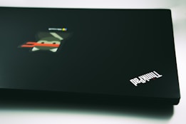 A black laptop featuring the ThinkPad logo in the lower right corner. The surface also includes a colorful sticker depicting a stylized character with a red band across its face, set against a blurred background.