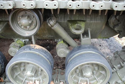 A close-up view of a tank's track system including the wheels, suspension components, and some parts of the tread. The metal appears to be weathered with patches of moss or dirt, and there are small fallen leaves at the base. The image highlights the intricate details and rugged construction of the track mechanism.