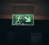 A glowing green emergency exit sign features a person walking through a door with an arrow pointing downwards and to the right. The sign is enclosed in a rectangular box suspended from the ceiling by two wires against a dark background.
